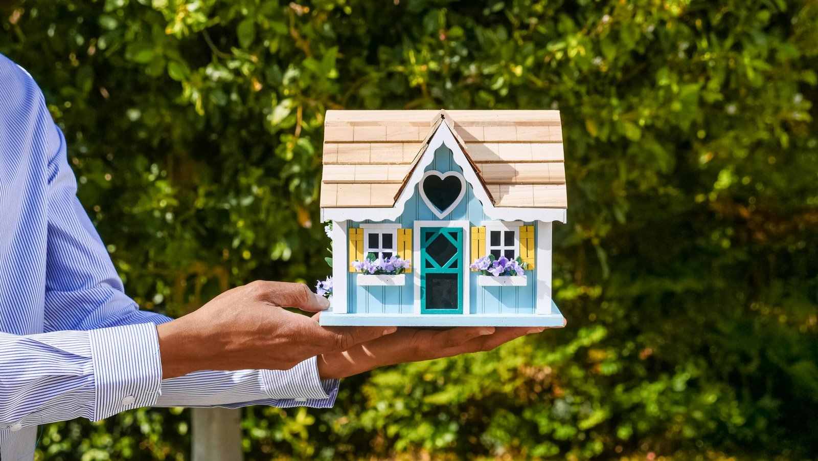 Close-up of a man's hands holding a decorative miniature house against foliage backdrop.